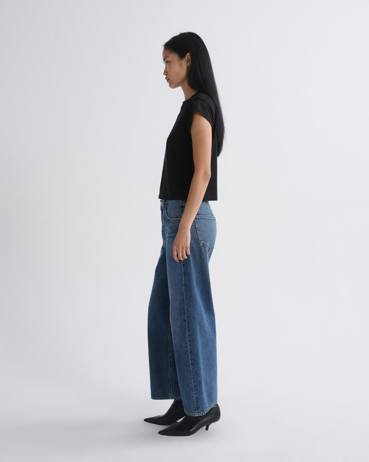 A model stands against a light gray studio background wearing a black short-sleeve top, blue wide-leg jeans, and black pointed ankle boots.