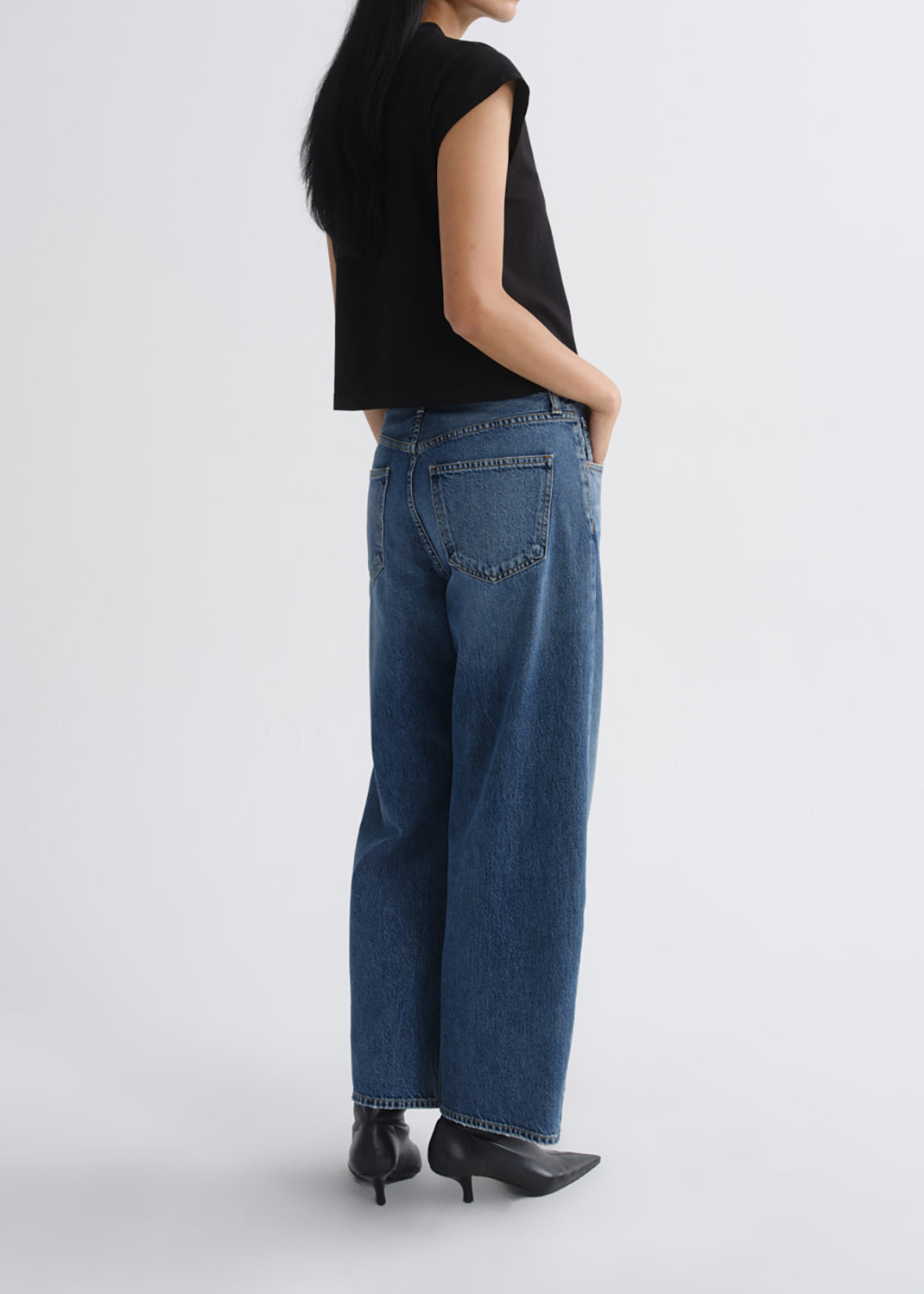A model shown from the back wearing blue wide-leg jeans, a black sleeveless top, and black pointed-toe heels against a grey background.