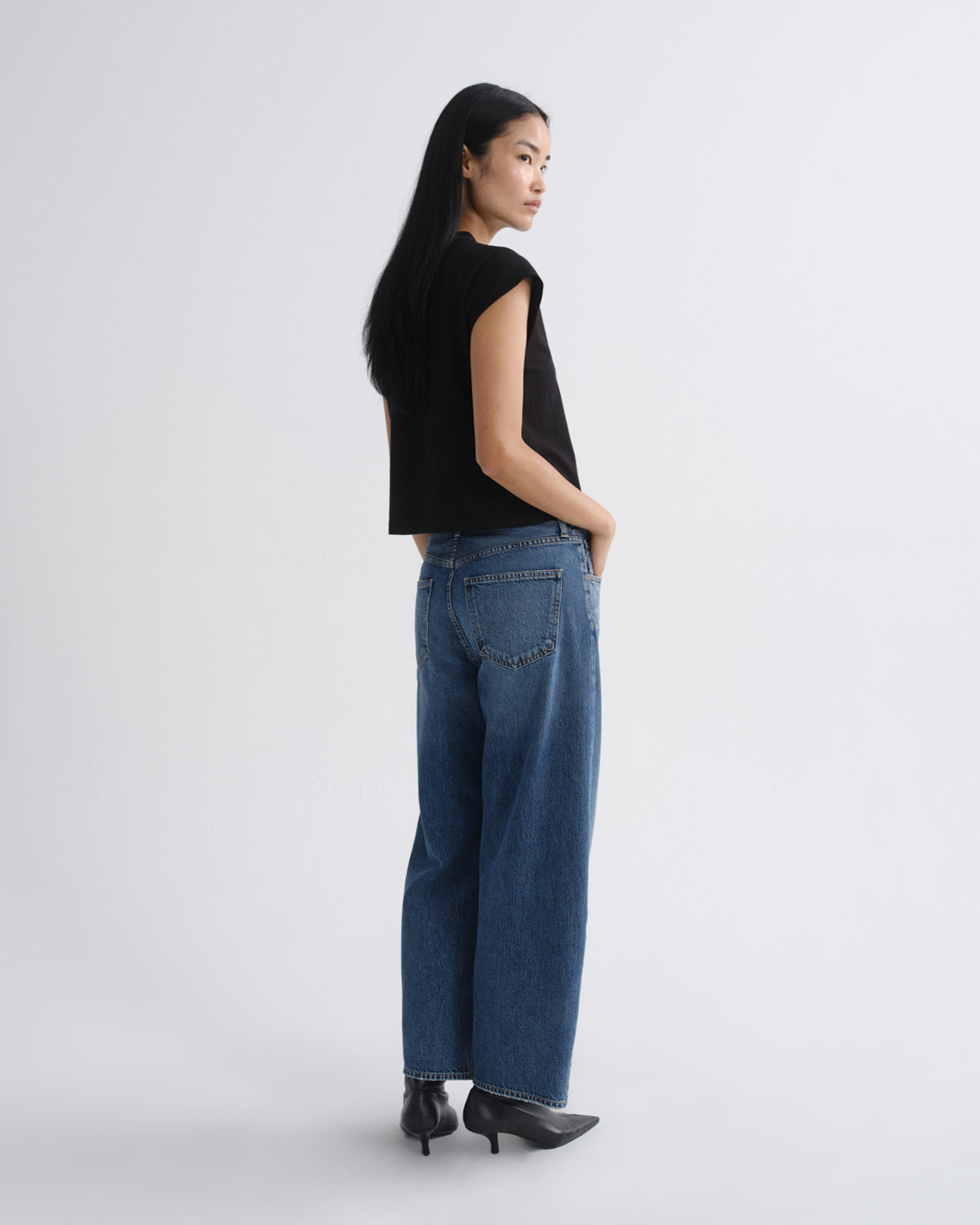 A model stands against a light gray studio background wearing a black short-sleeve top, blue wide-leg jeans, and black pointed ankle boots.