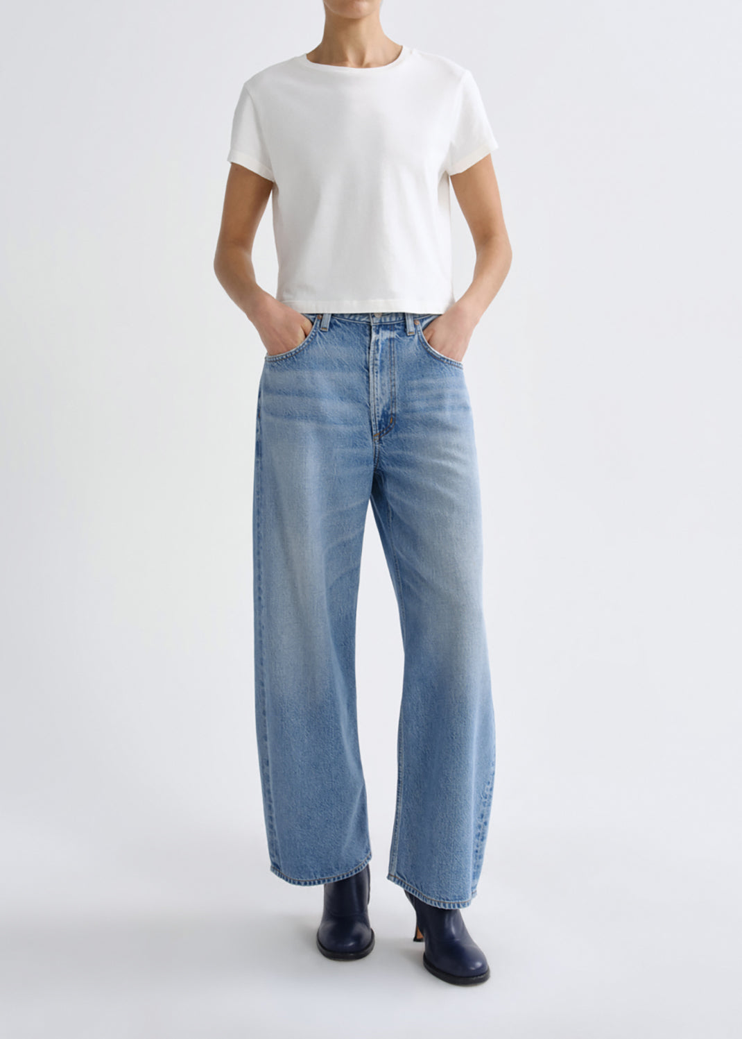 A model wearing a white short-sleeve t-shirt, light-wash wide-leg blue jeans, and black boots, standing against a grey background.