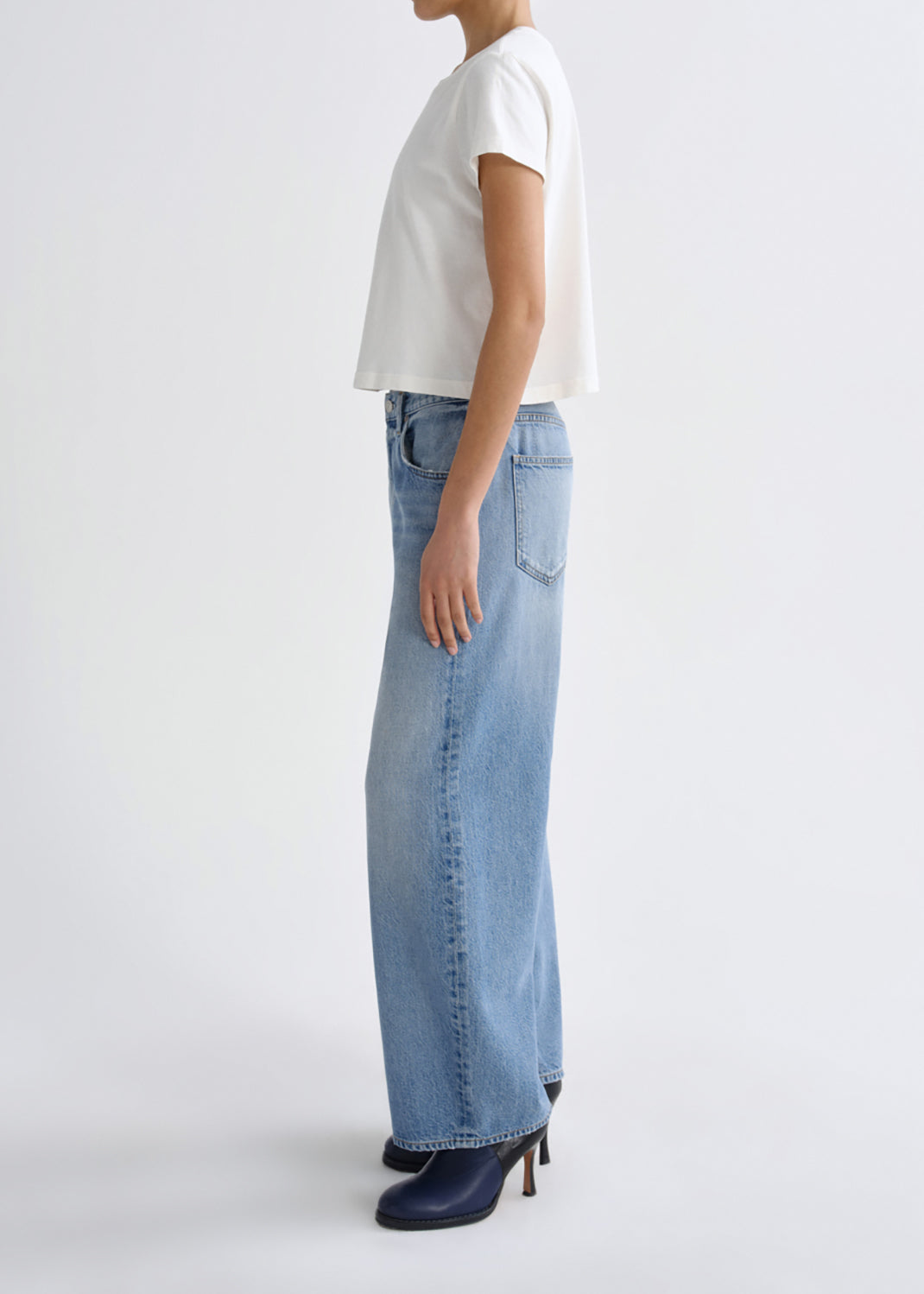 A model shown from the side wearing light-wash wide-leg denim jeans, a white short-sleeve top, and dark navy heeled boots against a grey background.