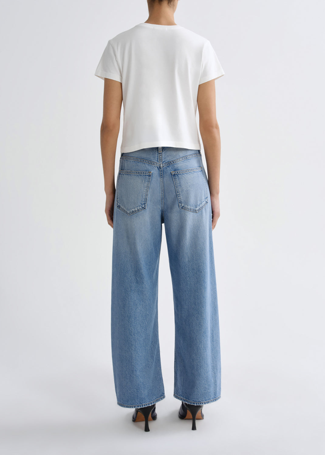 A model shown from the back wearing light-wash wide-leg denim jeans, a white short-sleeve crop top, and black high-heeled sandals against a grey background.