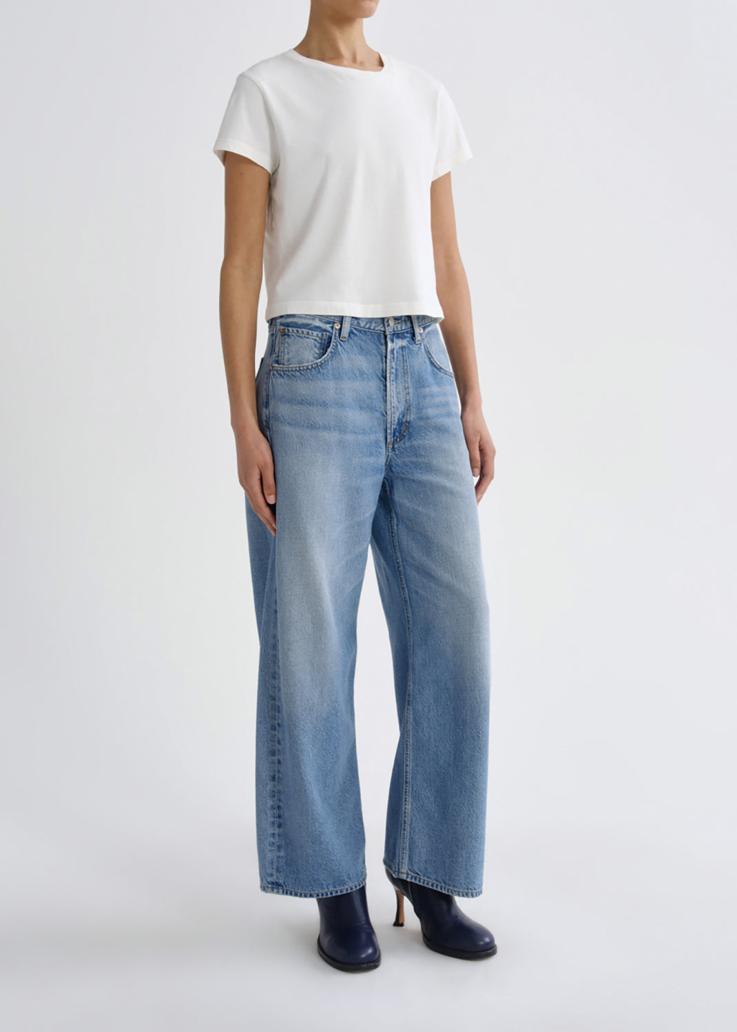 A model wearing light-wash wide-leg blue jeans, a white short-sleeve t-shirt, and dark navy boots, standing against a grey background.
