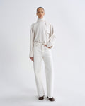 A model stands against a light gray studio background wearing a cream-colored long-sleeve mock-neck top with a draped collar detail and matching cream wide-leg trousers, paired with dark brown shoes.