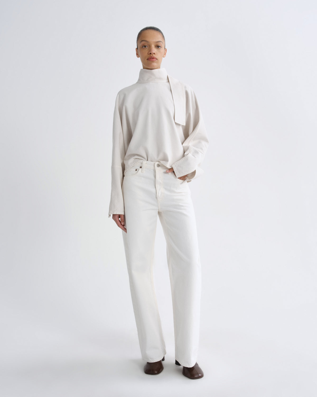 A model stands against a light gray studio background wearing a cream-colored long-sleeve mock-neck top with a draped collar detail and matching cream wide-leg trousers, paired with dark brown shoes.