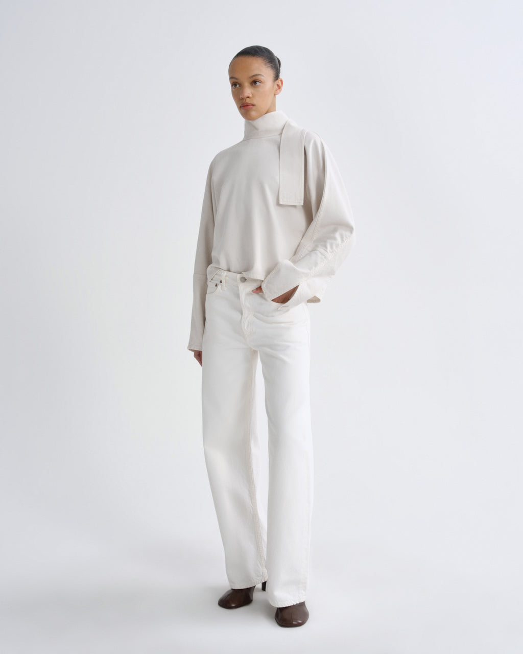A model stands against a seamless white studio background wearing a cream-colored long-sleeve mock-neck top with a wide collar detail and matching cream wide-leg trousers, paired with dark brown shoes.