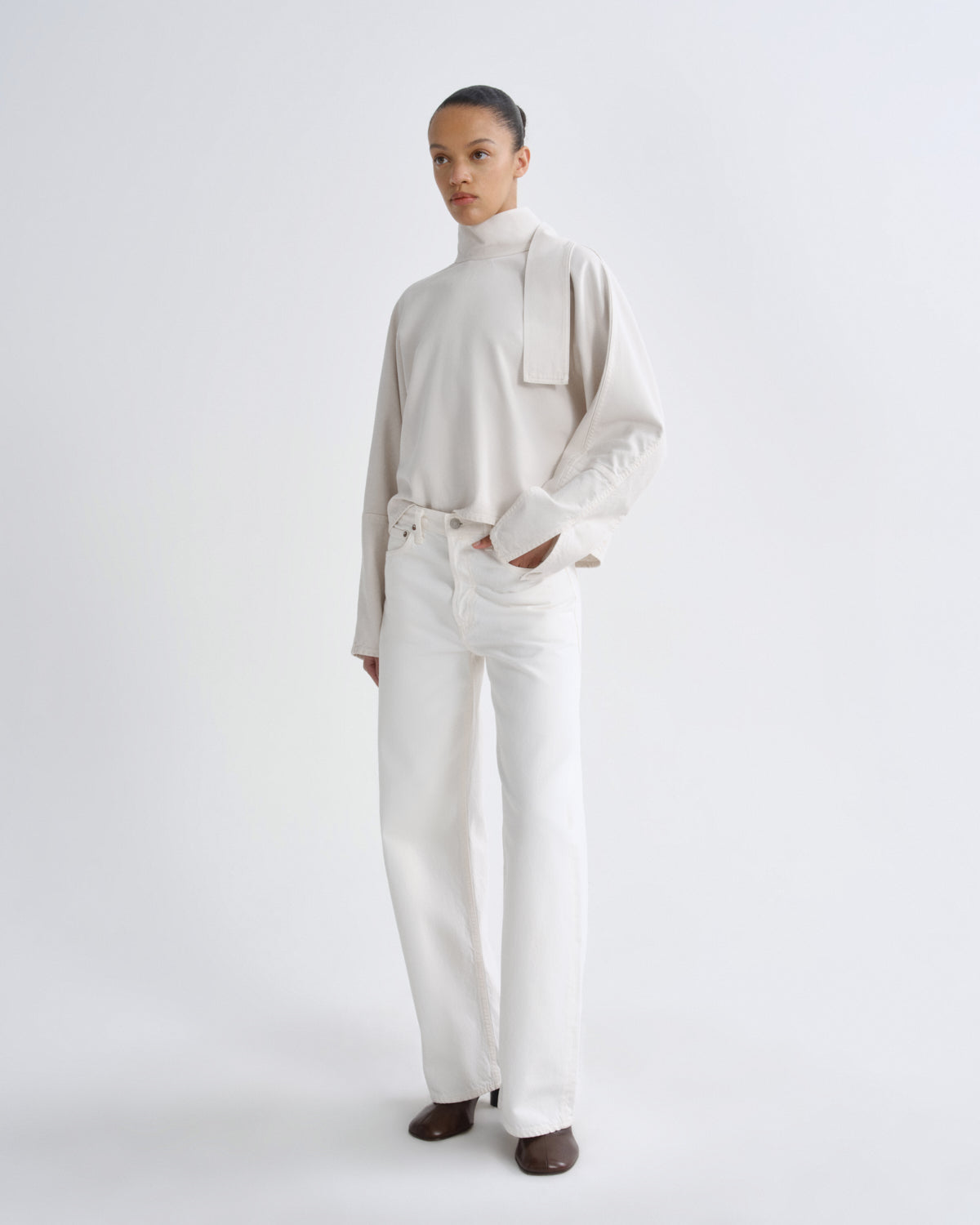 A model stands against a seamless white studio background wearing a cream-colored long-sleeve mock-neck top with a wide collar detail and matching cream wide-leg trousers, paired with dark brown shoes.