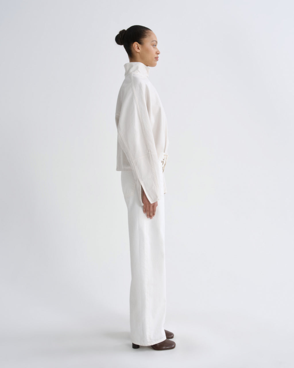 A model in profile wearing a white high-neck long-sleeve jacket and matching wide-leg trousers, standing against a seamless white studio background.