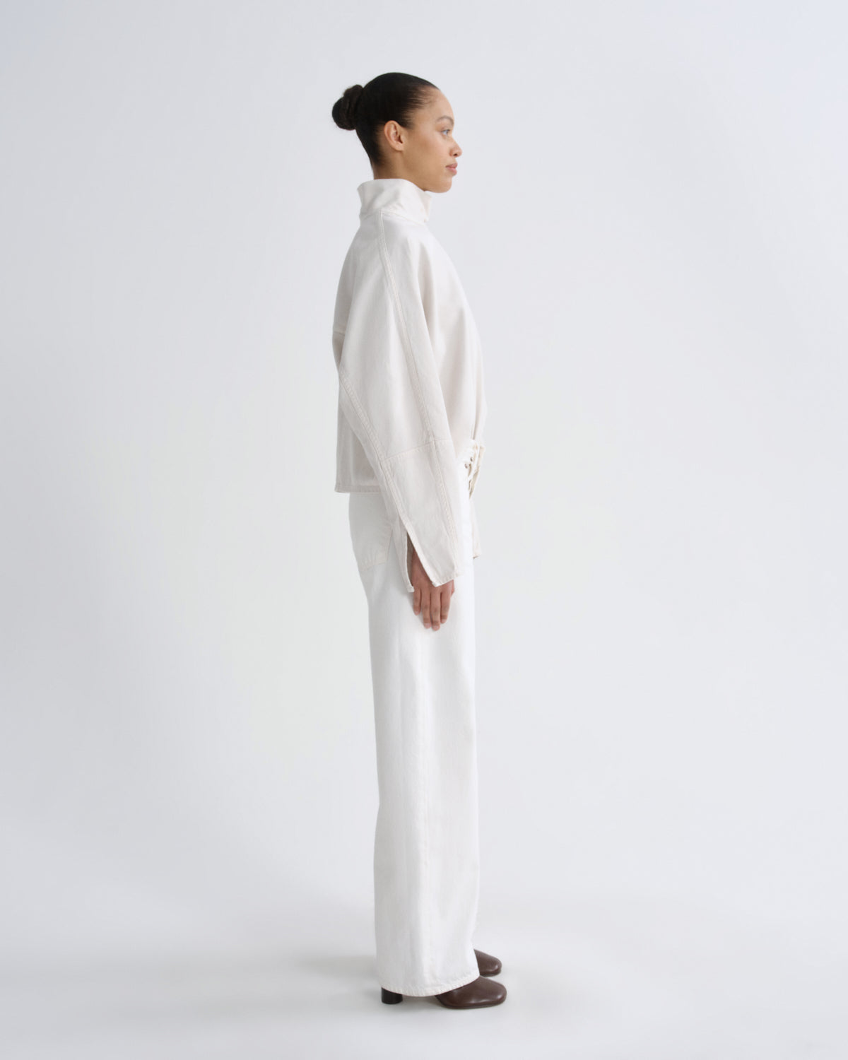 A model in profile wearing a white high-neck long-sleeve jacket and matching wide-leg trousers, standing against a seamless white studio background.