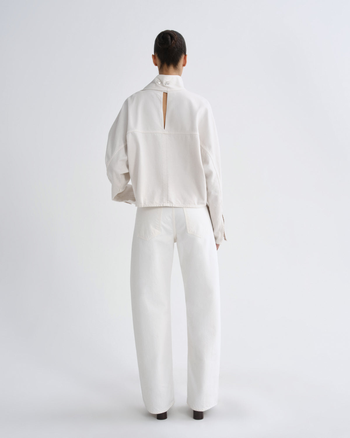 A model viewed from the back wearing a white long-sleeve cropped jacket with a high mock neck and a vertical slit at the back, paired with matching white wide-leg trousers, against a light gray studio background.
