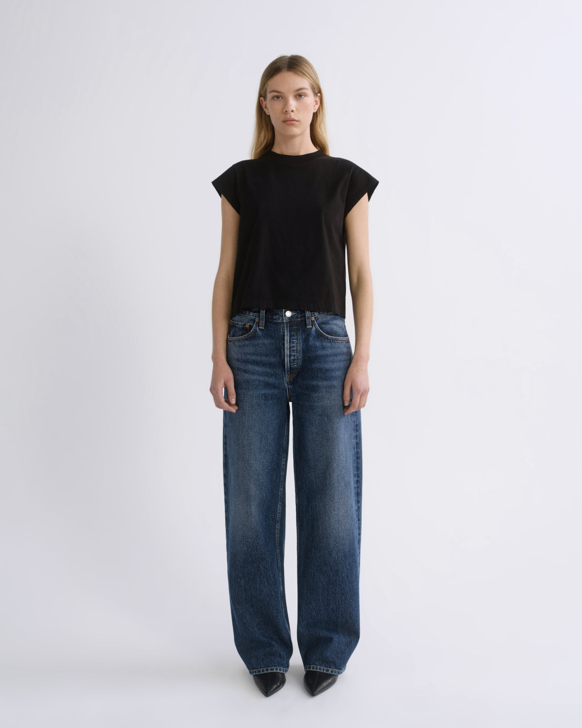 A model stands against a light gray studio background wearing a black short-sleeve crewneck top and dark blue wide-leg jeans.