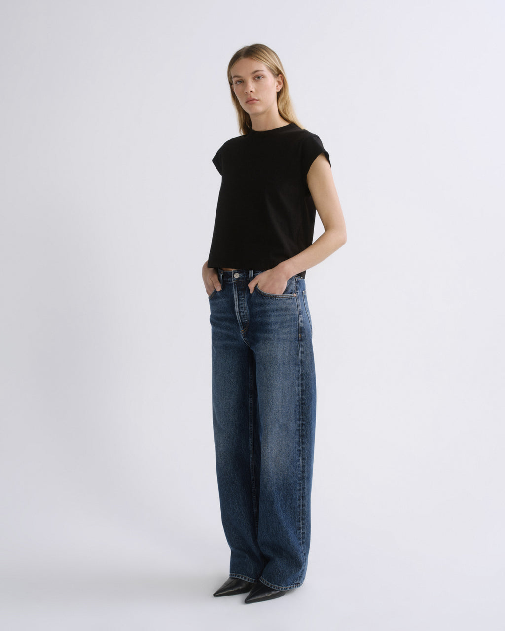 A woman modeling a black short-sleeve crewneck top and wide-leg blue jeans against a plain light gray studio background.