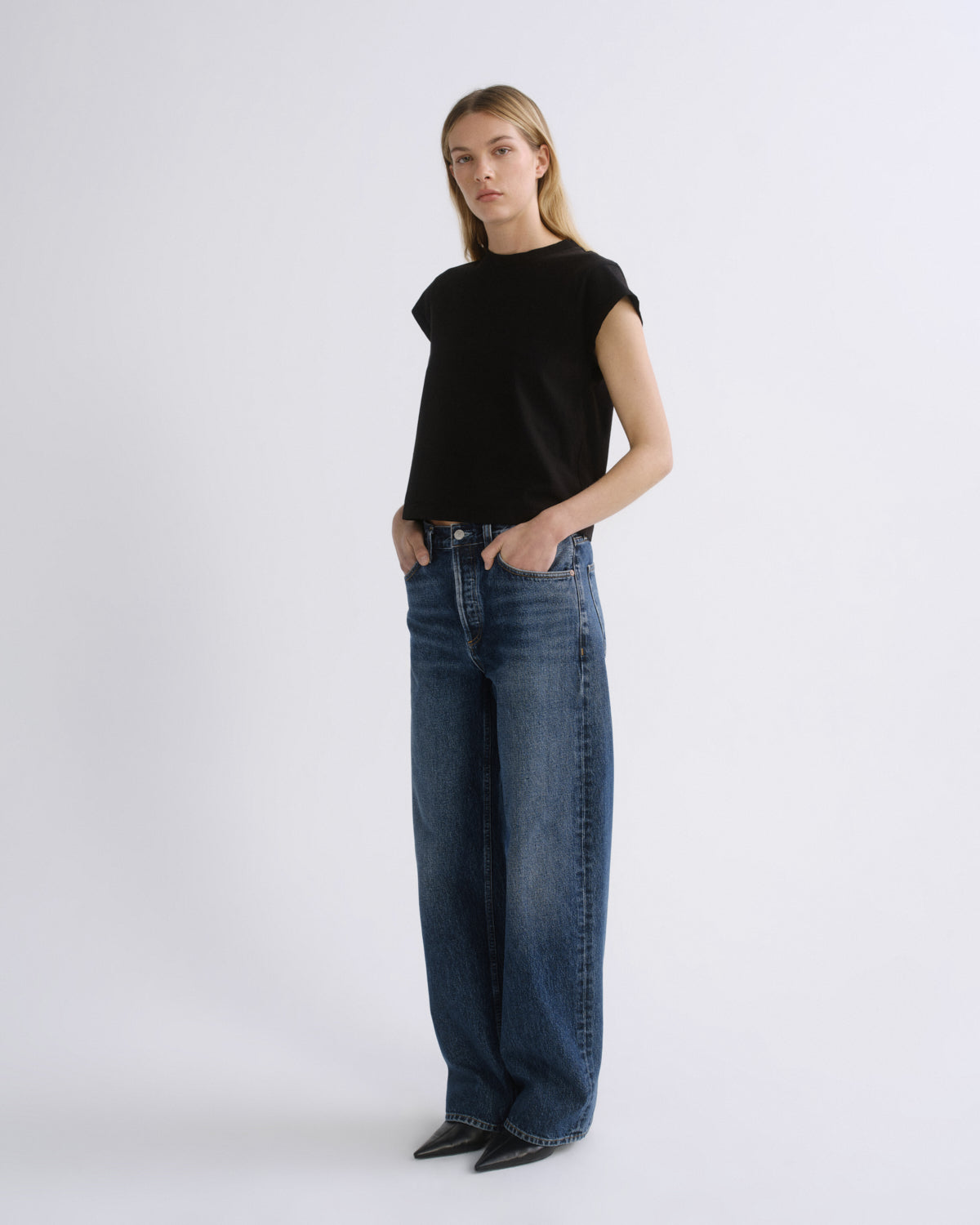A woman modeling a black short-sleeve crewneck top and wide-leg blue jeans against a plain light gray studio background.