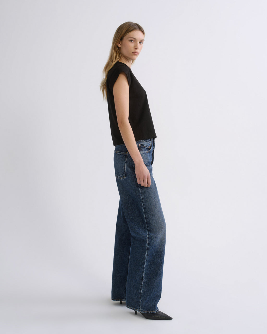 A woman stands in profile wearing a black sleeveless top and wide-leg blue jeans against a plain light gray studio background.