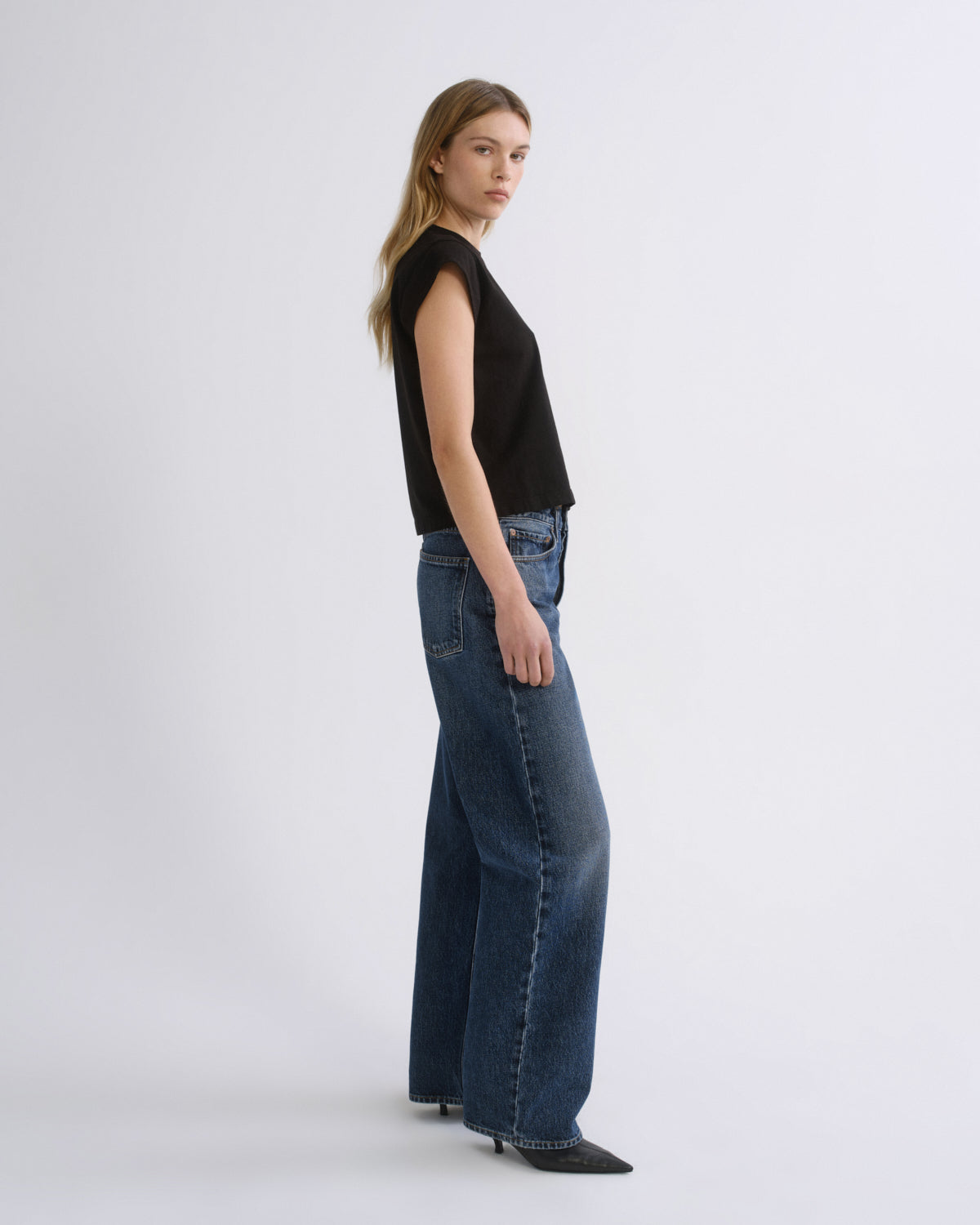 A woman stands in profile wearing a black sleeveless top and wide-leg blue jeans against a plain light gray studio background.