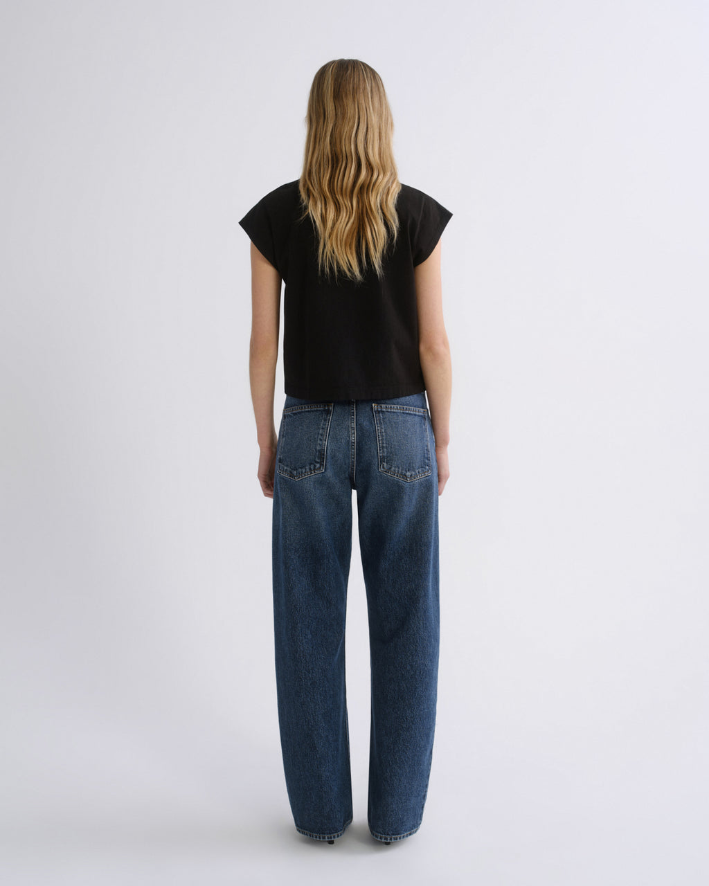 A woman viewed from behind wearing a black short-sleeve t-shirt and wide-leg blue jeans, standing against a plain light gray studio background.