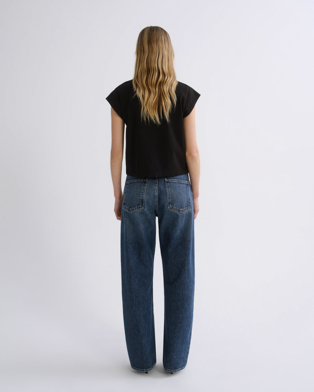 A woman viewed from behind wearing a black short-sleeve t-shirt and wide-leg blue jeans, standing against a plain light gray studio background.