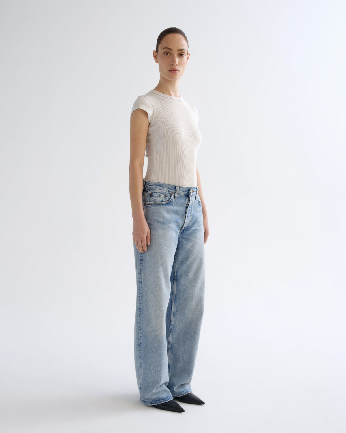 A model stands in profile wearing a fitted off-white short-sleeve top and light-wash wide-leg blue jeans against a clean white studio background.