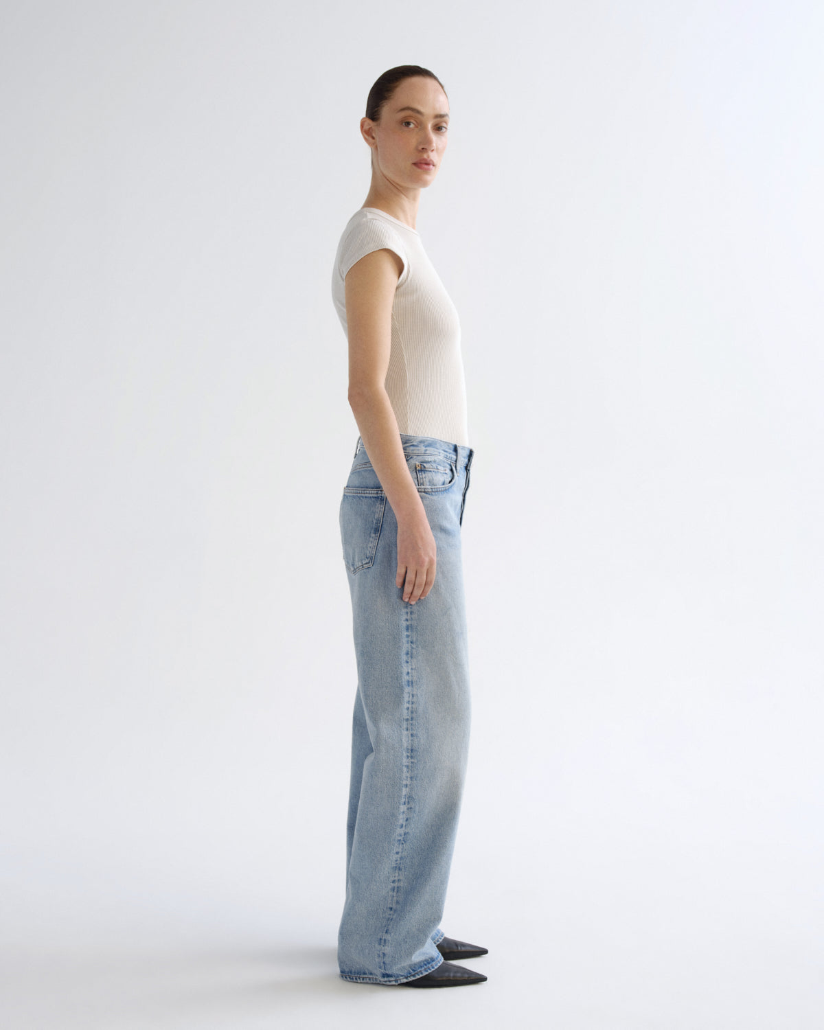 A model standing in profile wearing a fitted off-white short-sleeve top and light-wash wide-leg blue jeans against a plain light gray studio background.