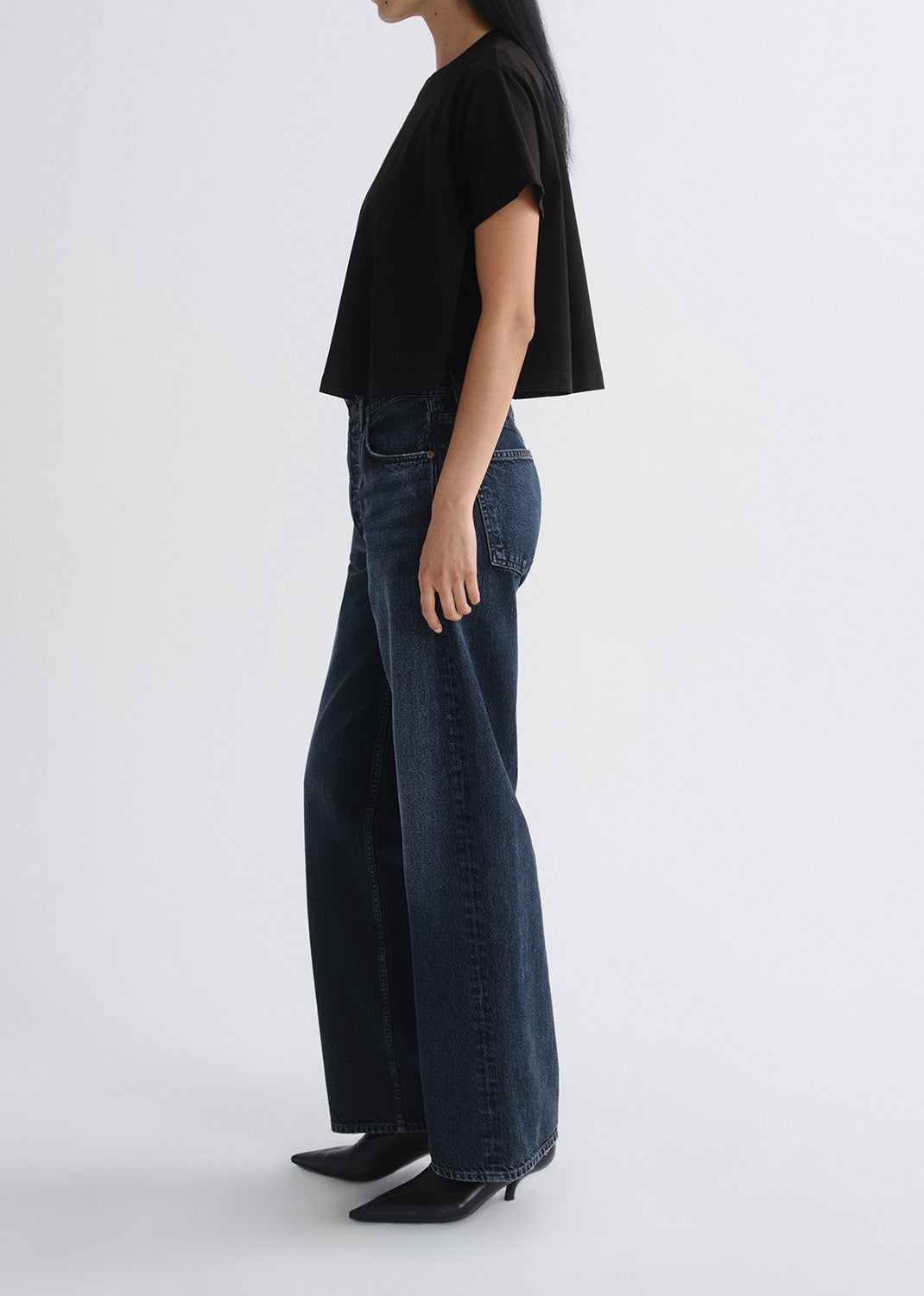 Person wearing a black short sleeve crewneck tee with dark wash wide leg jeans viewed from the side on a grey background