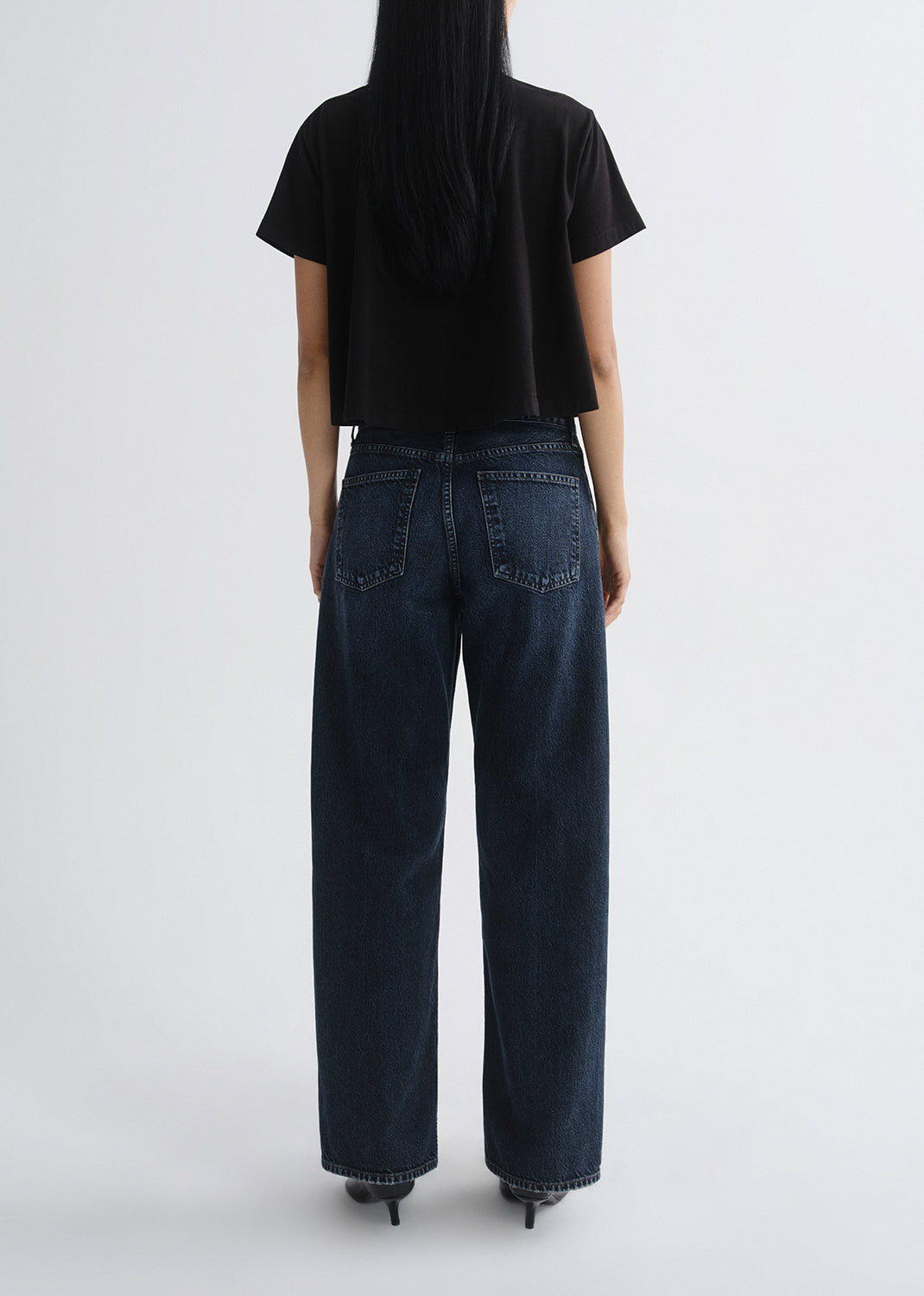Person wearing a black short sleeve crewneck tee with dark wash wide leg jeans viewed from the back on a grey background