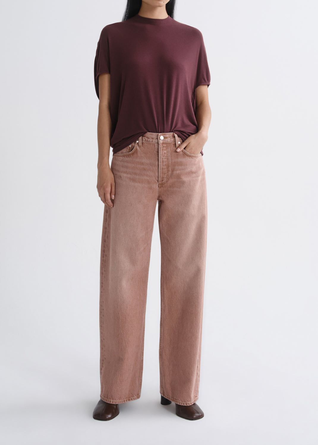 A model wearing a dark burgundy short-sleeve mock neck top and dusty rose wide-leg jeans, standing against a grey background.