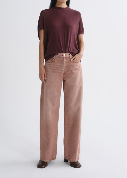 A model wearing a dark burgundy short-sleeve mock neck top and dusty rose wide-leg jeans, standing against a grey background.