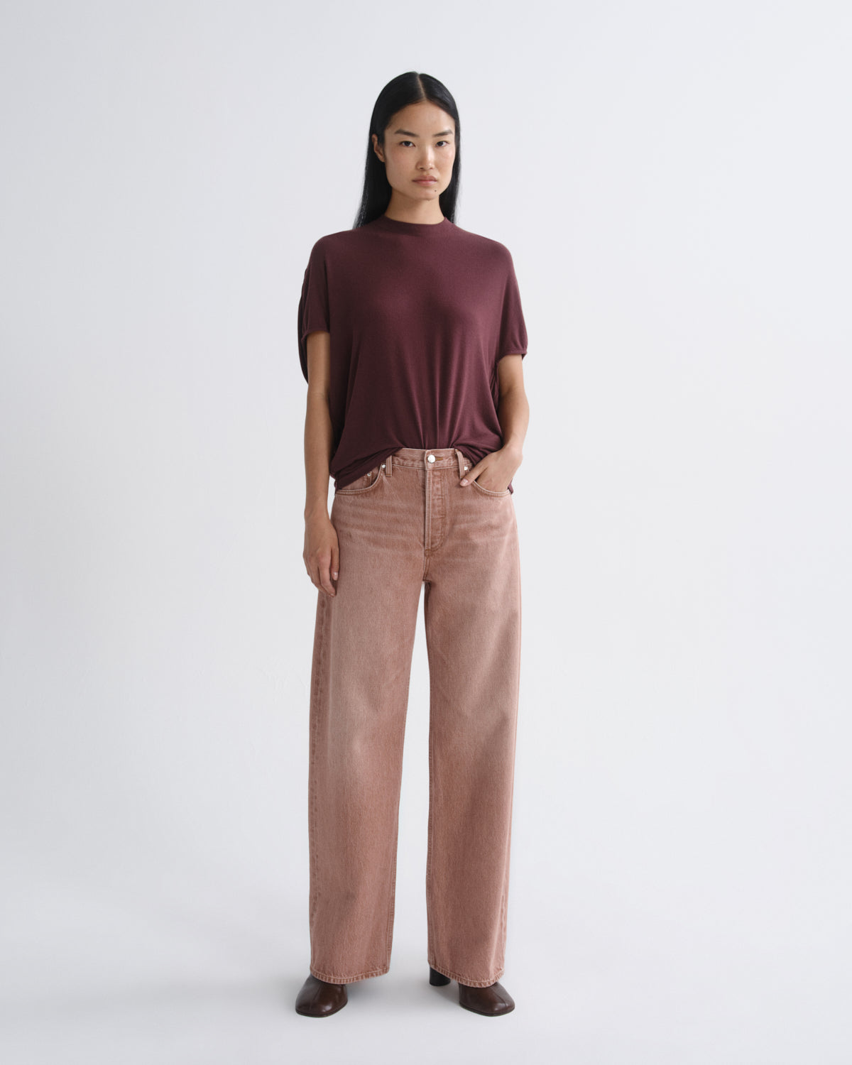 A model stands against a light gray studio background wearing a short-sleeve crewneck t-shirt in deep burgundy and high-waisted, wide-leg corduroy pants in a dusty rose pink.