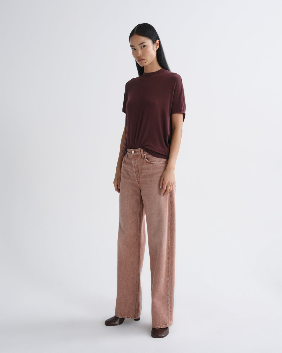 A model stands against a light gray studio background wearing a dark burgundy short-sleeve t-shirt and dusty rose wide-leg jeans.