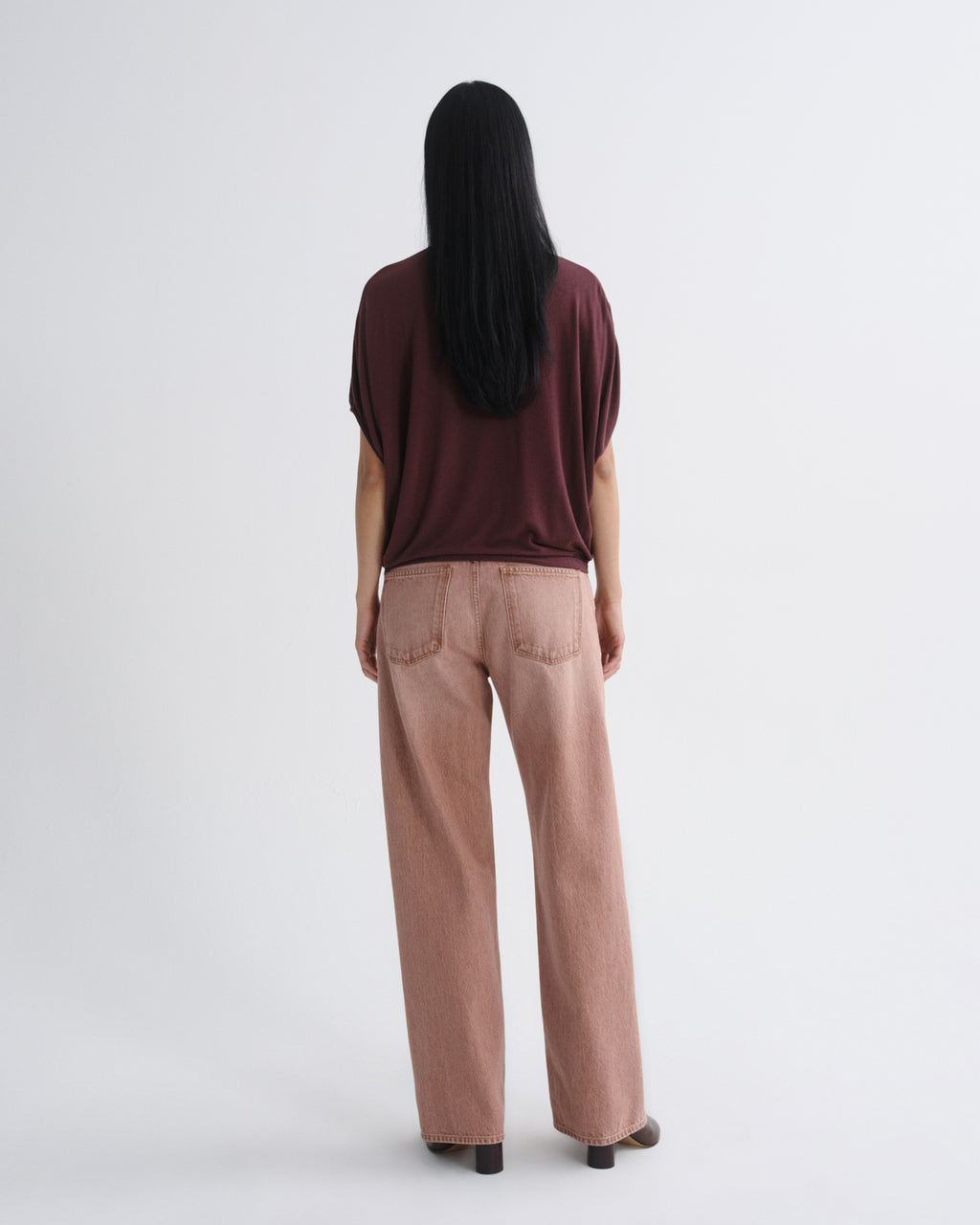 A woman viewed from behind wearing a dark burgundy short-sleeve top and dusty rose wide-leg jeans against a plain light gray studio background.