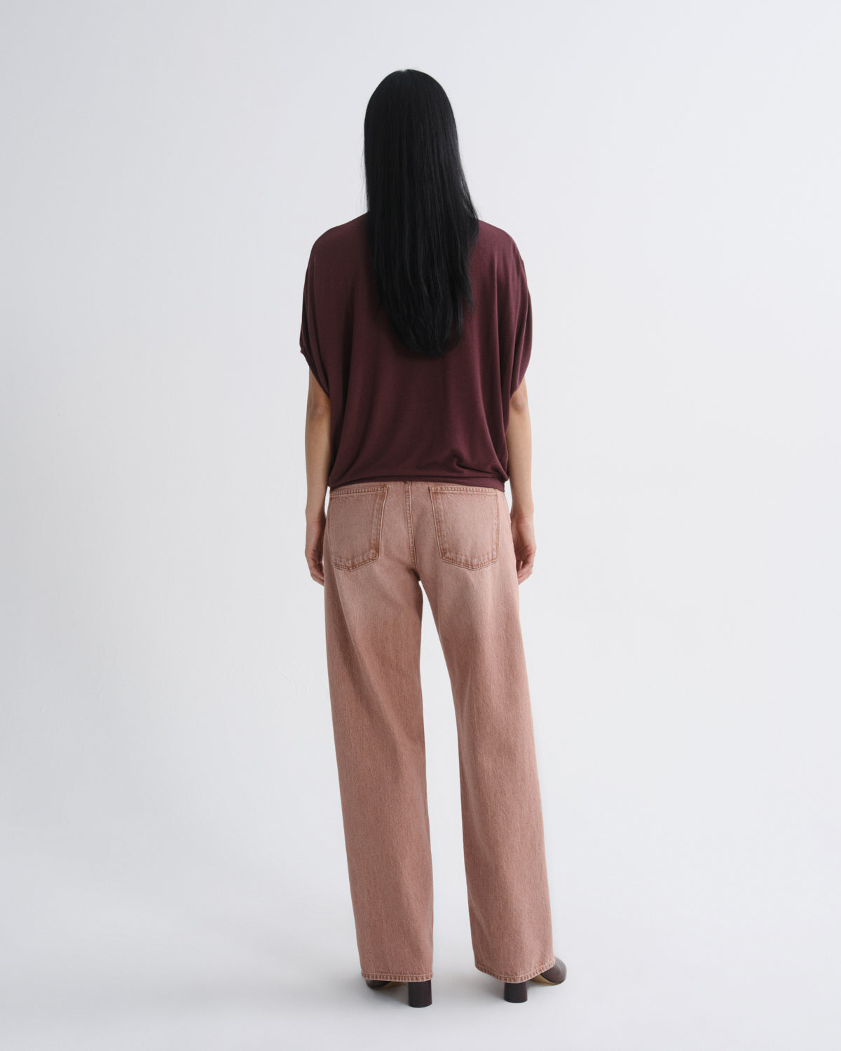 A woman viewed from behind wearing a dark burgundy short-sleeve top and dusty rose wide-leg jeans against a plain light gray studio background.