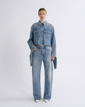 A model stands against a light gray studio background wearing a light blue cropped denim jacket with matching wide-leg jeans, a cream turtleneck, and white pointed shoes.