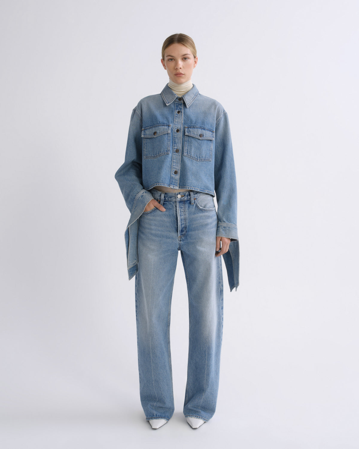 A model stands against a light gray studio background wearing a light blue cropped denim jacket with matching wide-leg jeans, a cream turtleneck, and white pointed shoes.