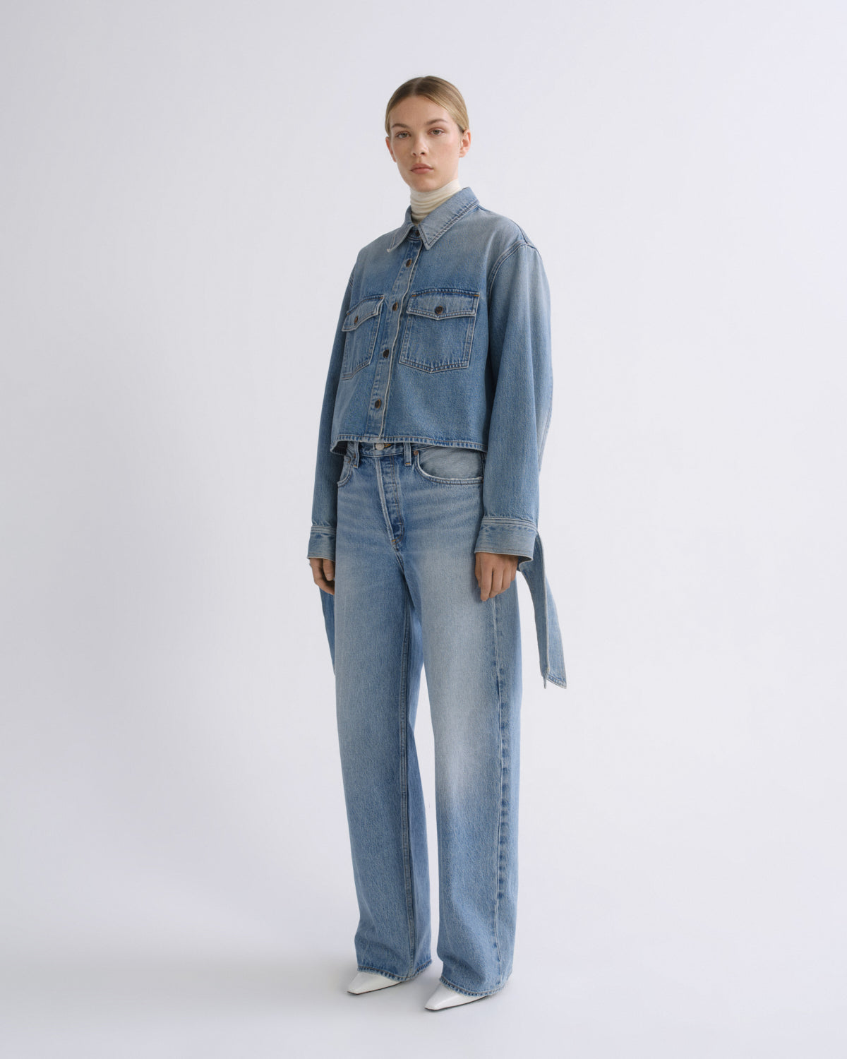 A model stands against a light gray studio background wearing a light blue denim jacket with matching wide-leg jeans, a white turtleneck, and white pointed shoes.