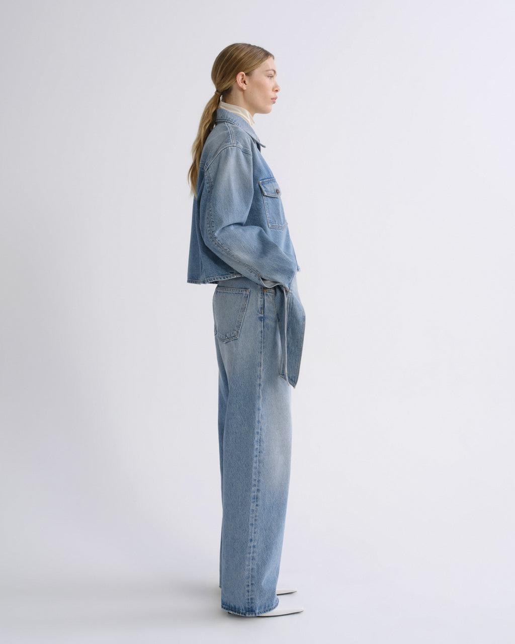 A model in side profile wearing a light blue denim jacket and matching wide-leg jeans, standing against a clean white studio background.