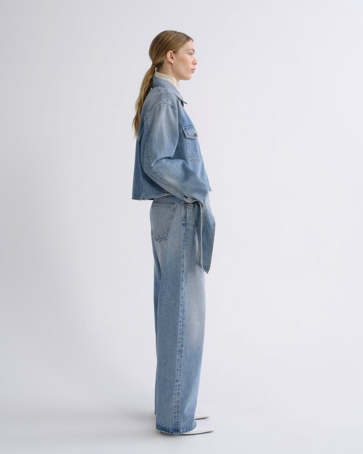 A model in side profile wearing a light blue denim jacket and matching wide-leg jeans, standing against a clean white studio background.