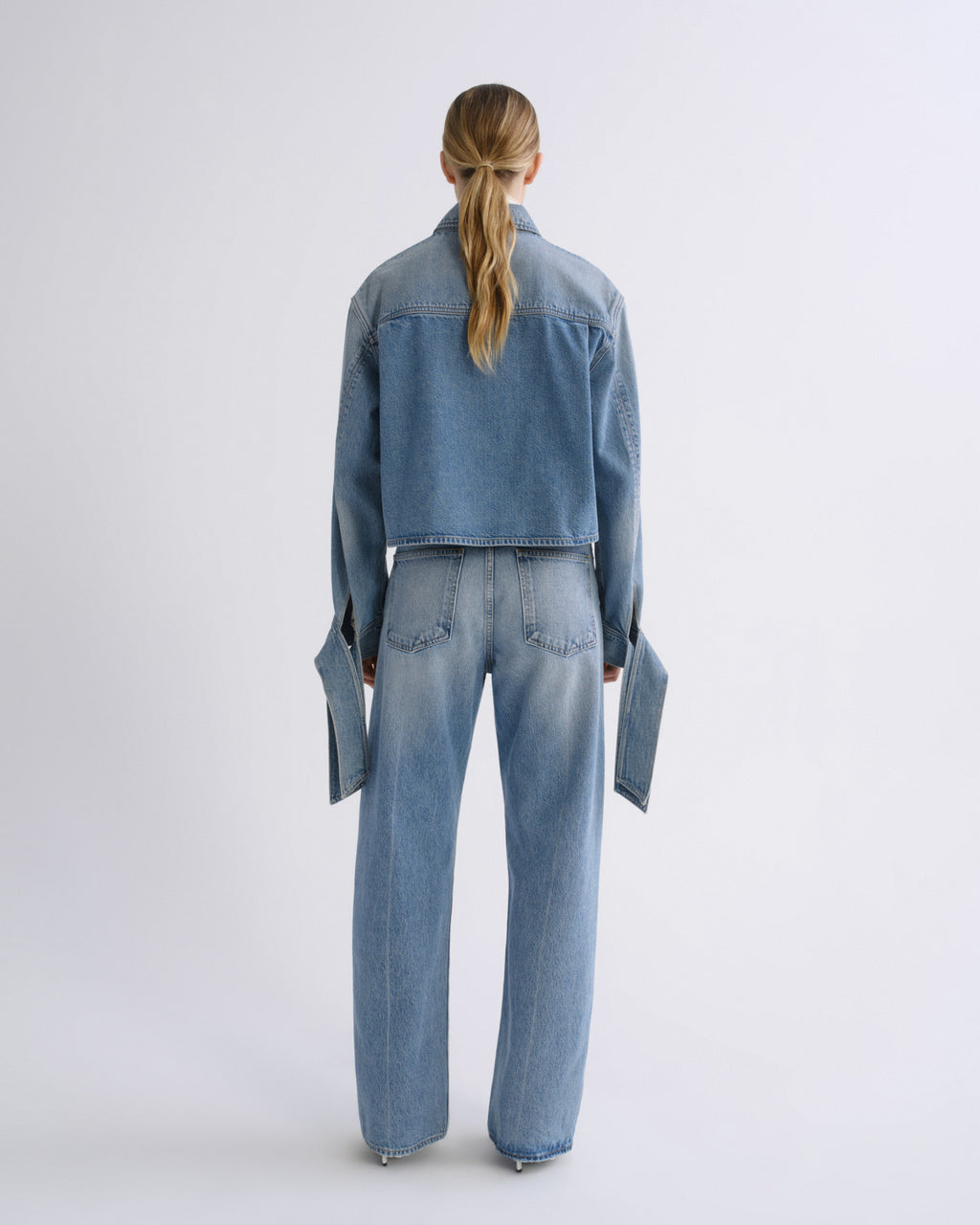 A model viewed from the back wearing a light blue denim jacket and matching wide-leg jeans against a plain light gray studio background.