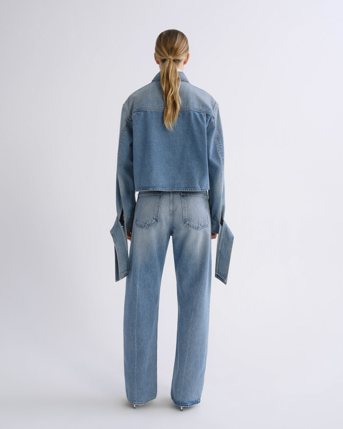 A model viewed from the back wearing a light blue denim jacket and matching wide-leg jeans against a plain light gray studio background.