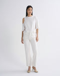 A model stands against a light gray studio background wearing a white cold-shoulder top with elbow-length sleeves, matching white wide-leg cropped pants, and beige heeled sandals.