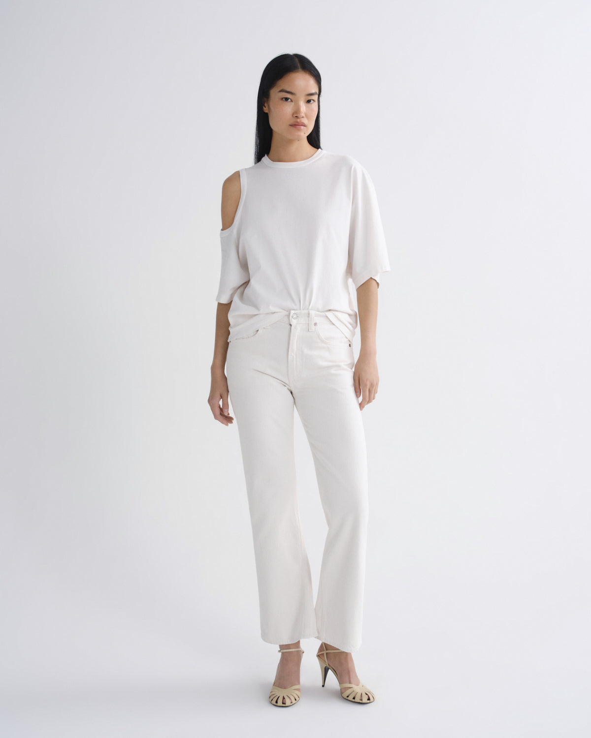 A model stands against a light gray studio background wearing a white cold-shoulder top with elbow-length sleeves, matching white wide-leg cropped pants, and beige heeled sandals.