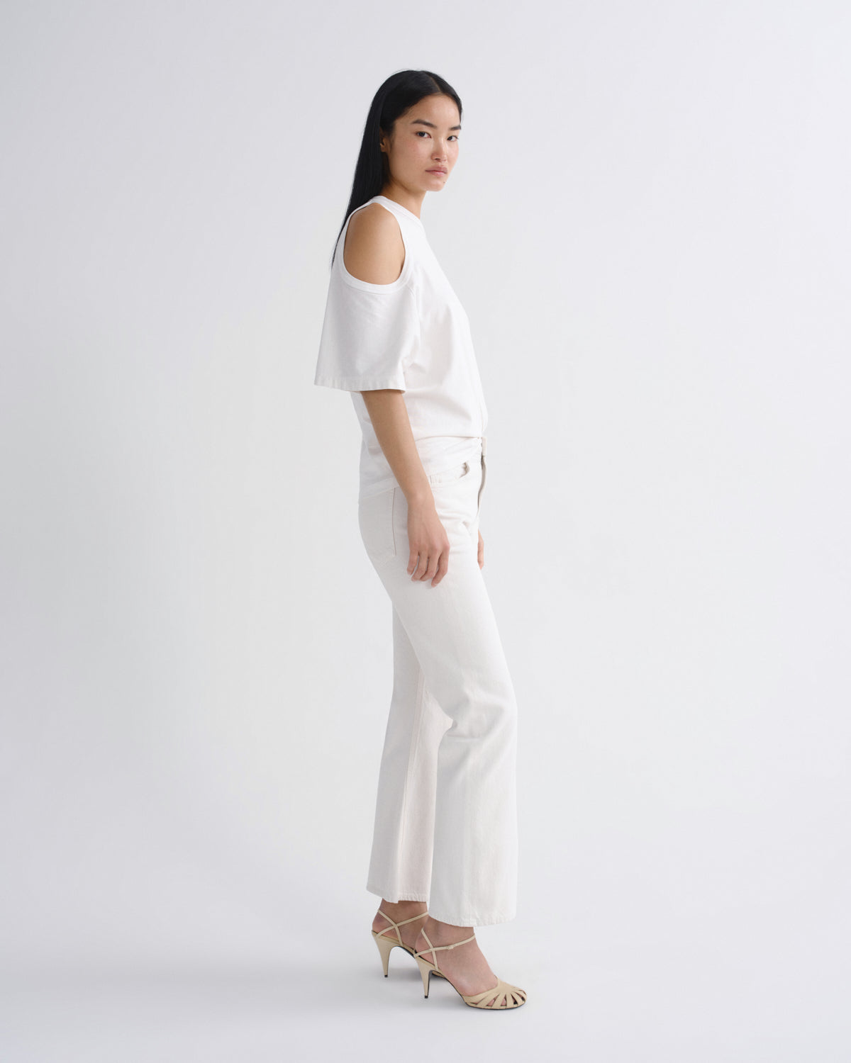 A model wearing a white cold-shoulder top and matching white wide-leg trousers, paired with beige strappy high-heeled sandals, standing in profile against a light gray studio background.