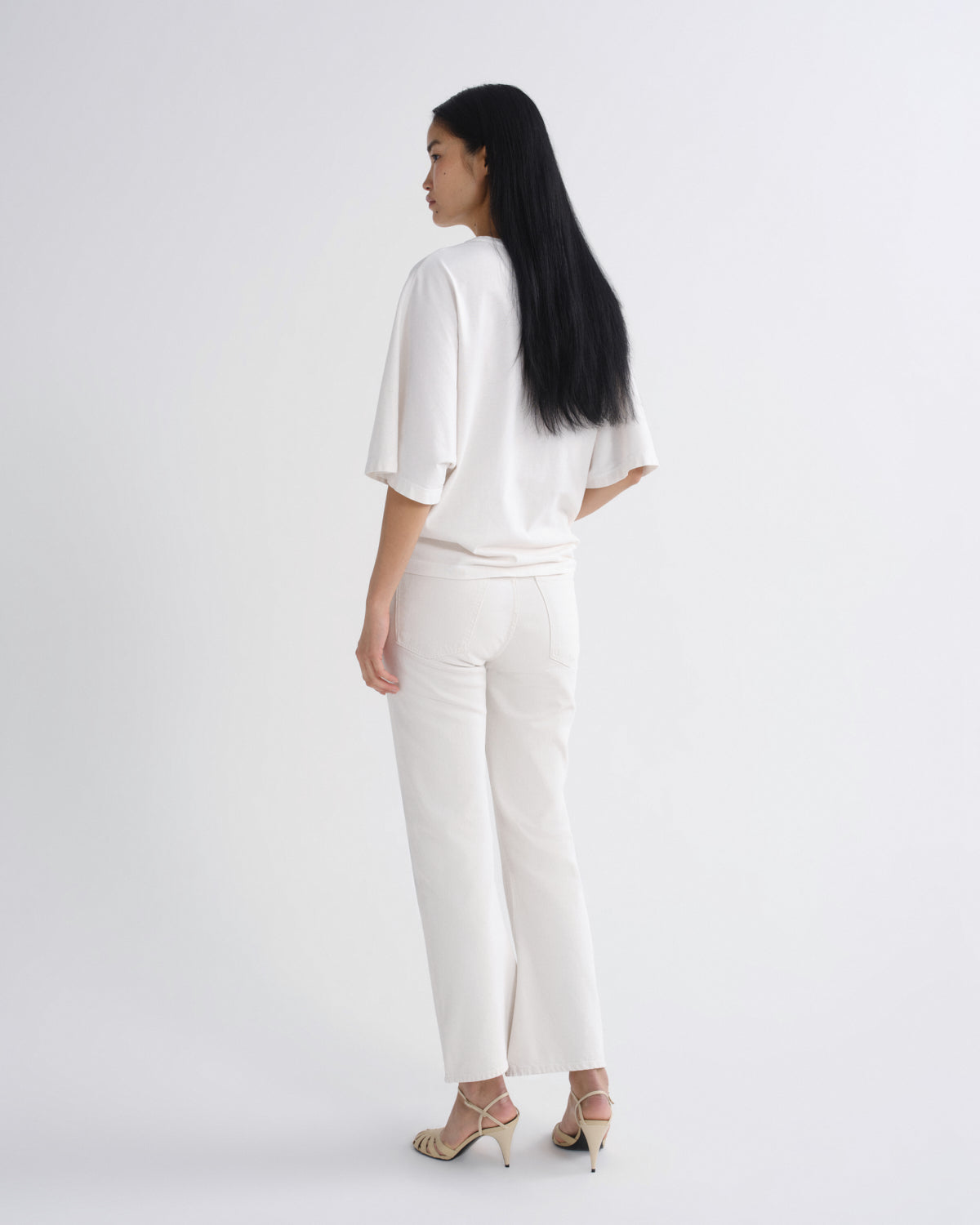 A woman with long black hair is shown from the back wearing a matching white two-piece set consisting of a short-sleeve top and wide-leg trousers, paired with beige high-heeled sandals, against a plain white studio background.
