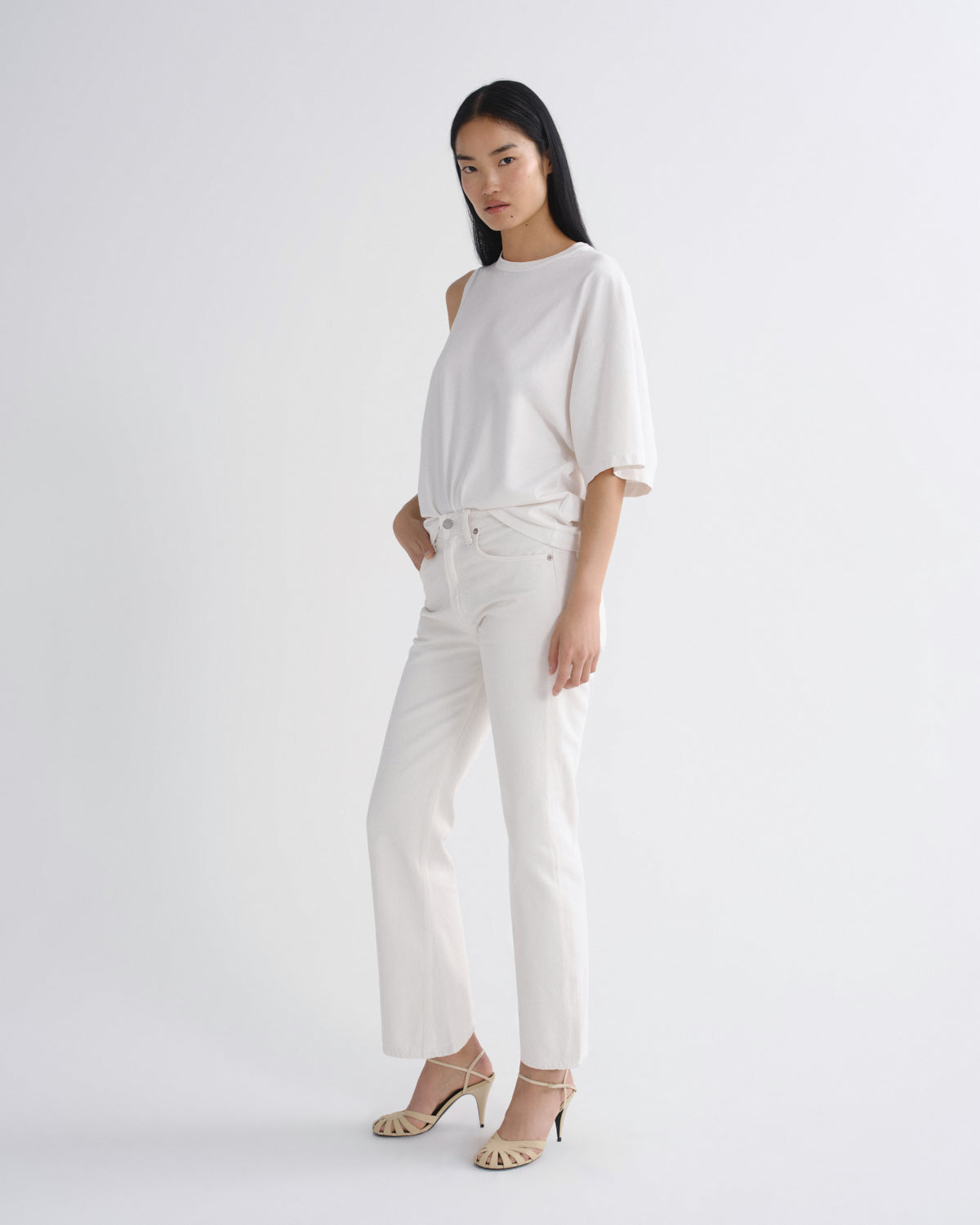 A model wearing a white short-sleeve top and white wide-leg trousers, paired with beige high-heeled sandals, standing against a light gray studio background.