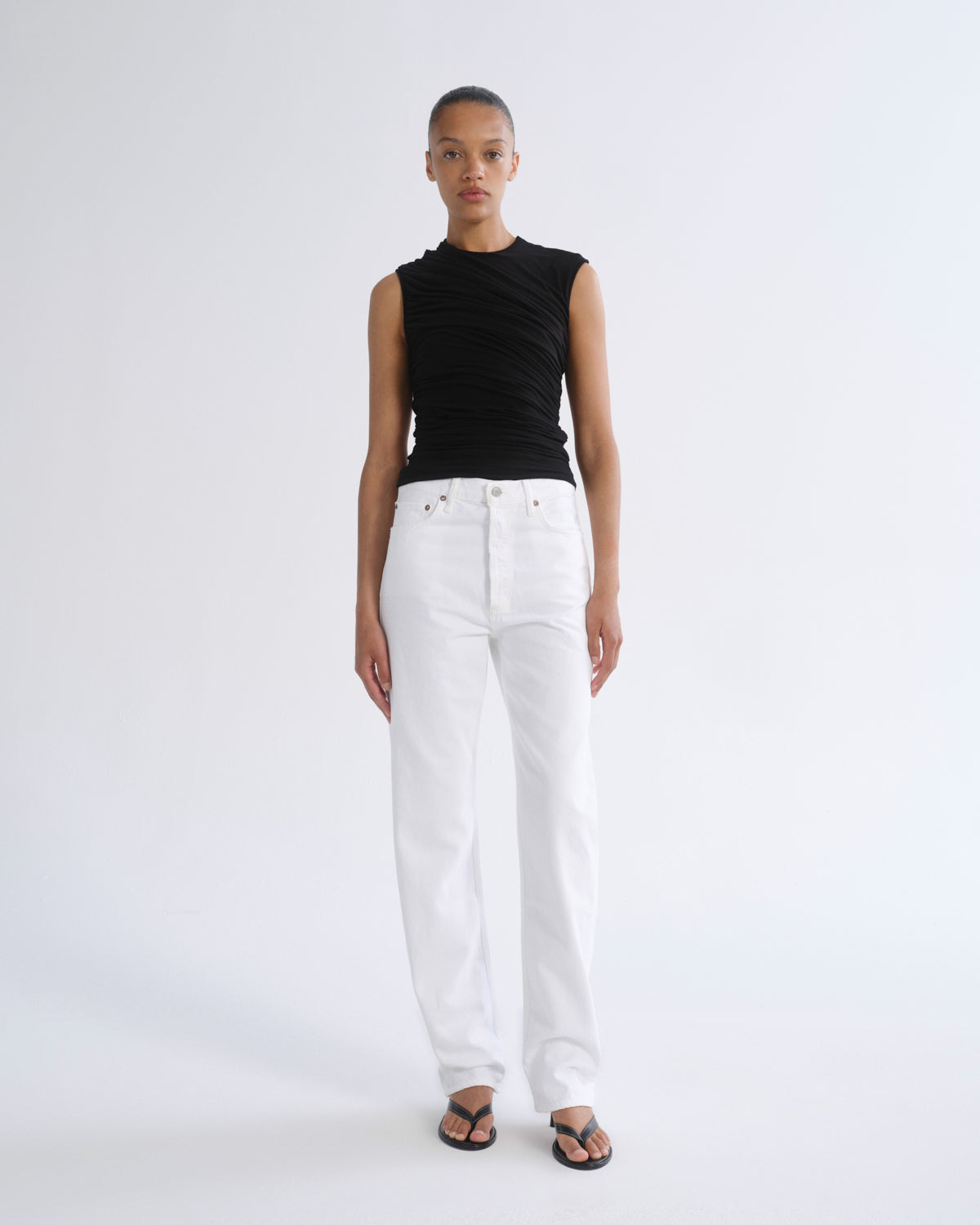 A model stands against a light gray studio background wearing a sleeveless black top, white straight-leg jeans, and black sandals.