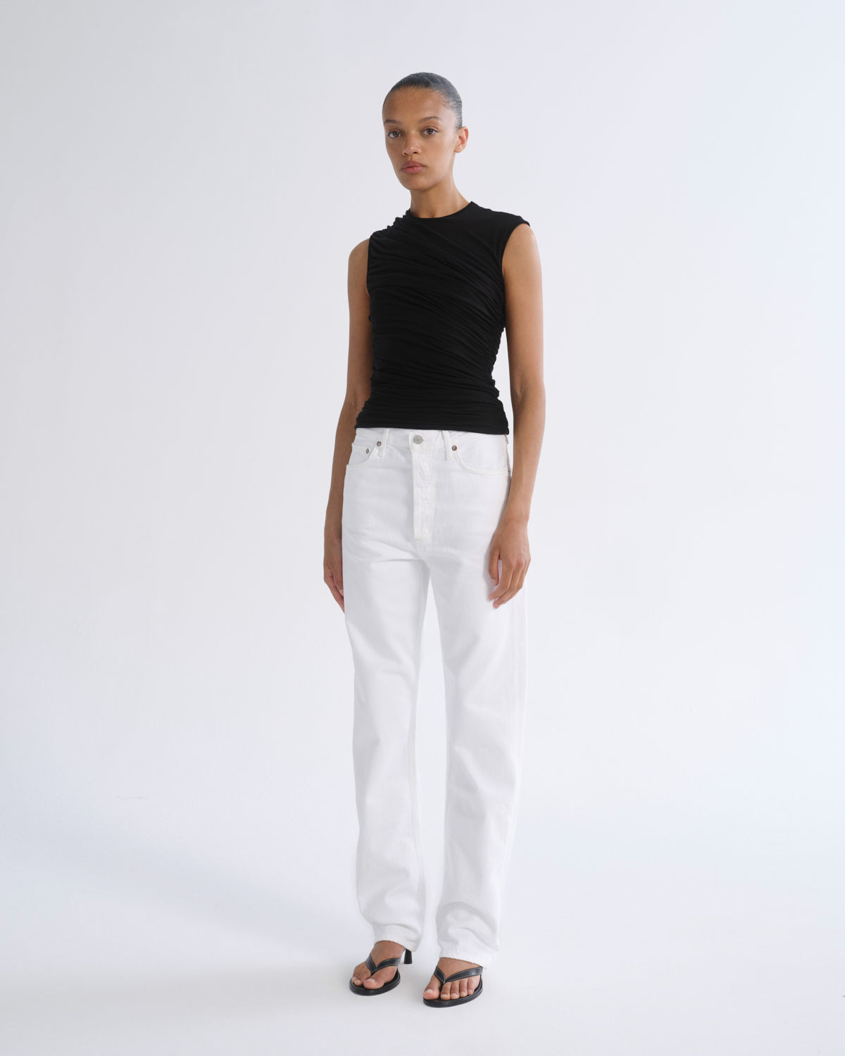 A model stands against a light gray studio background wearing a sleeveless black ruched top, white straight-leg jeans, and black sandals.