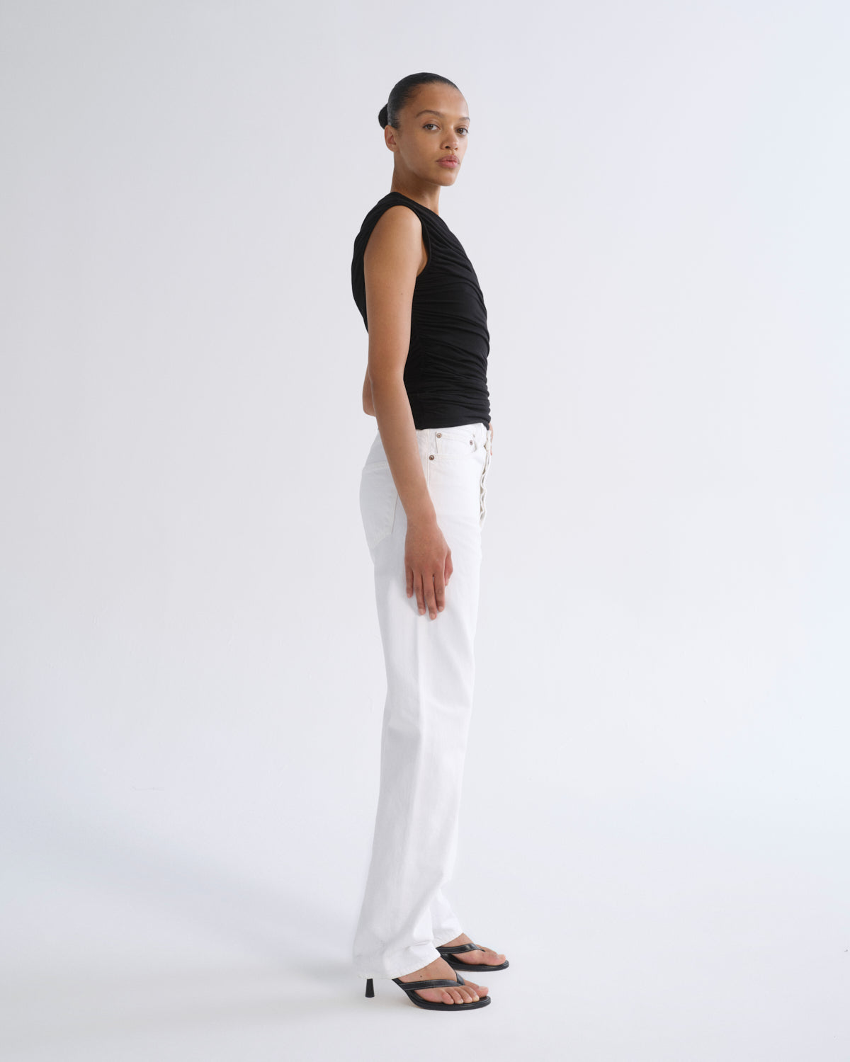 A model stands in profile wearing a black sleeveless ribbed top, white wide-leg pants with gold-tone hardware, and black sandals against a plain light gray studio background.