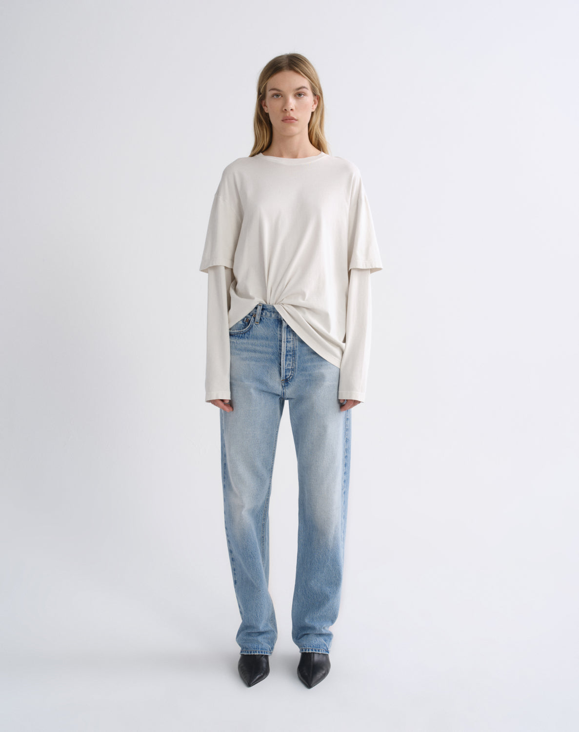 A model stands against a light gray studio background wearing a cream-colored, long-sleeve knit top with a twisted hem detail, paired with light-wash blue jeans and black pointed-toe heels.
