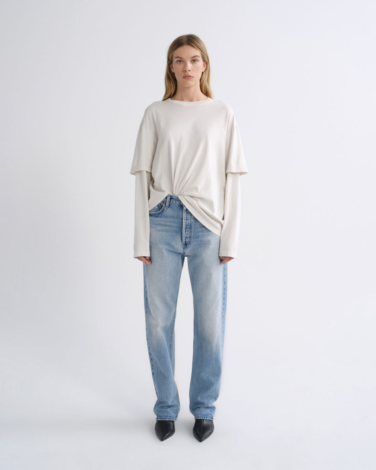A model stands against a light gray studio background wearing a cream-colored, long-sleeve knit top with a twisted hem detail, paired with light-wash blue jeans and black pointed-toe heels.