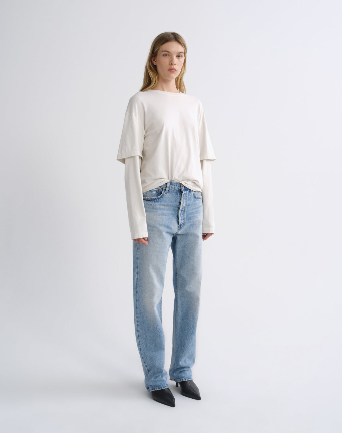 A model stands against a light gray studio background wearing a cream-colored long-sleeve top with layered sleeves, light-wash blue jeans, and black pointed-toe heels.
