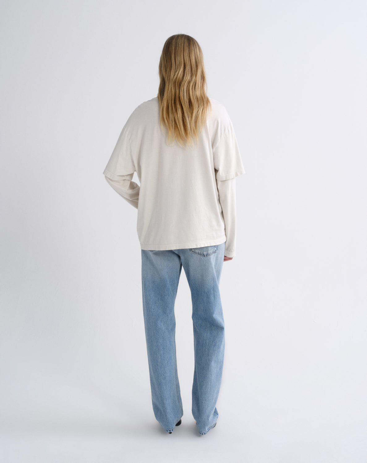 A woman viewed from behind wearing a cream-colored long-sleeve top and light blue wide-leg jeans, standing against a plain light gray studio background.